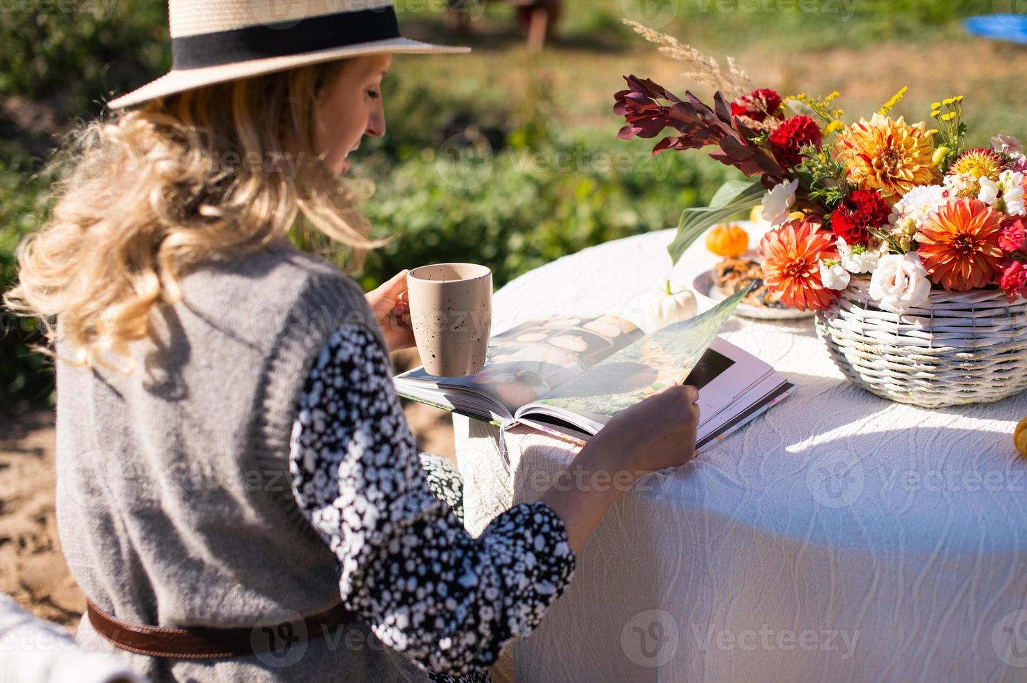 un mujer voltea mediante un libro y bebidas un caliente bebida a un mesa con flores en el calle foto