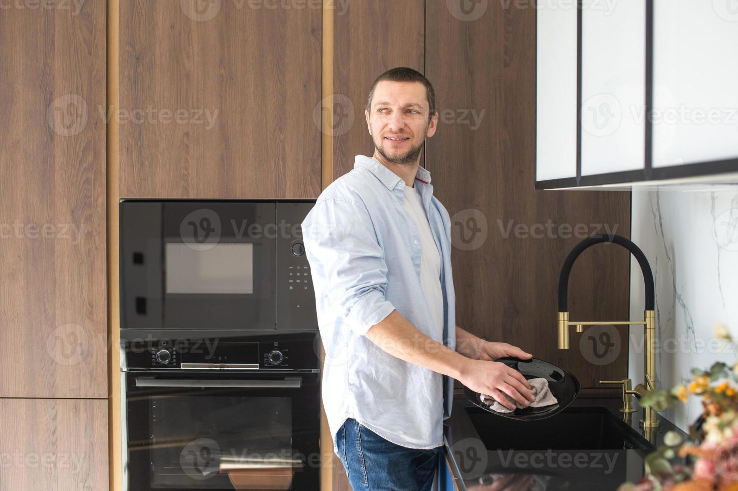 un hombre en un camisa toallitas platos con un toalla en pie en el cocina. casa quehaceres foto