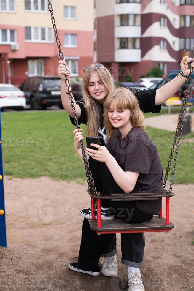 Two funny girls are sitting on a swing in the city and taking selfies on their phone. Teenagers smile photo