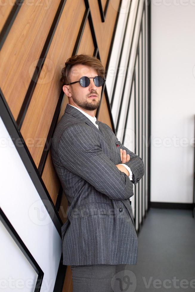 a young man in a suit and sunglasses is standing in the office near the wall photo