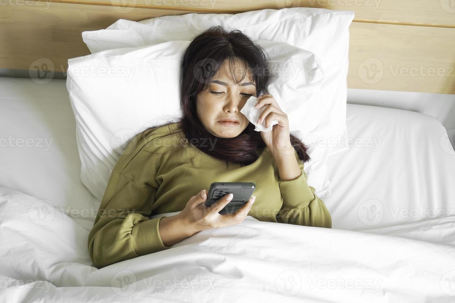 A sad young Asian woman is crying over her phone while lying on the bed photo