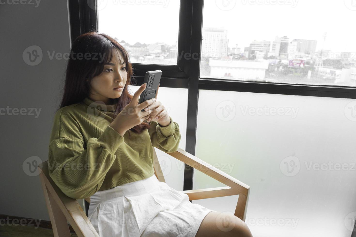 A young Asian woman is feeling serious and focused on holding her smartphone in hand while sitting in a bedroom. photo