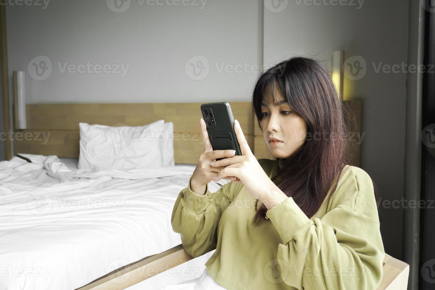 A young Asian woman is feeling serious and focused on holding her smartphone in hand while sitting in a bedroom. photo