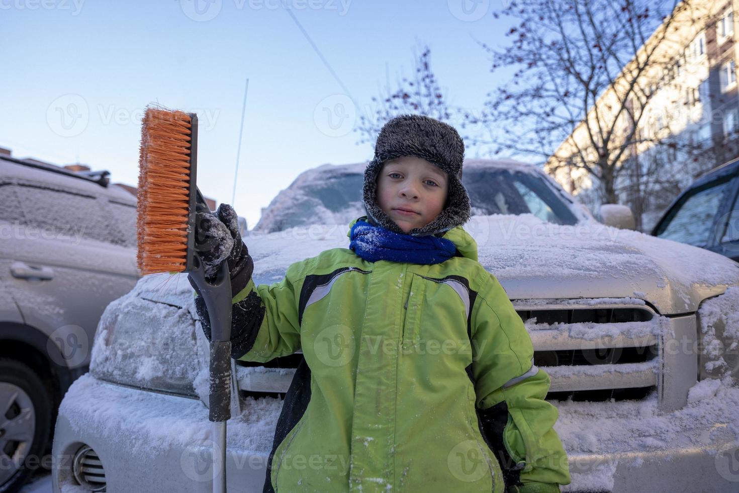 Funny cute Caucasian boy cleans the car from snow with a brush and a scraper photo