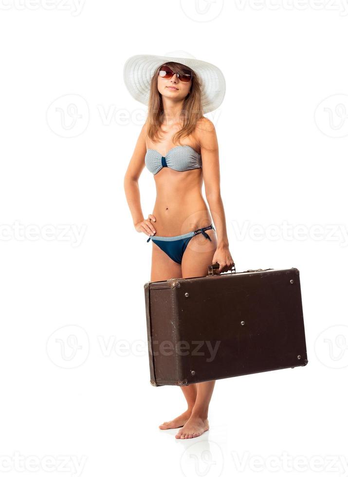 Full length portrait of a beautiful young woman posing in a bikini, hat and sunglasses with a suitcase in hand on white photo
