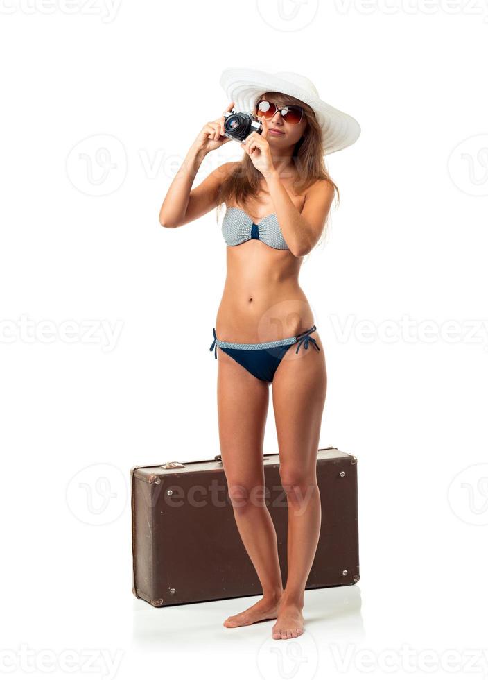 Full length portrait of a beautiful young woman posing in a bikini with a camera in hands and suitcase on white photo