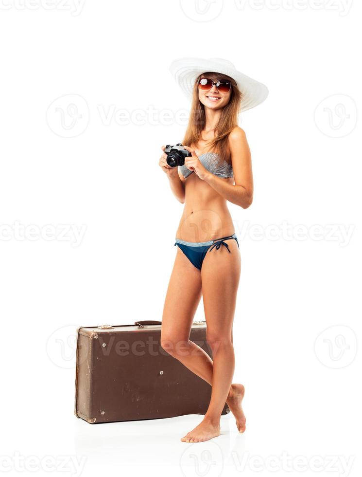 Full length portrait of a beautiful young woman posing in a bikini with a camera in hands and suitcase on white photo