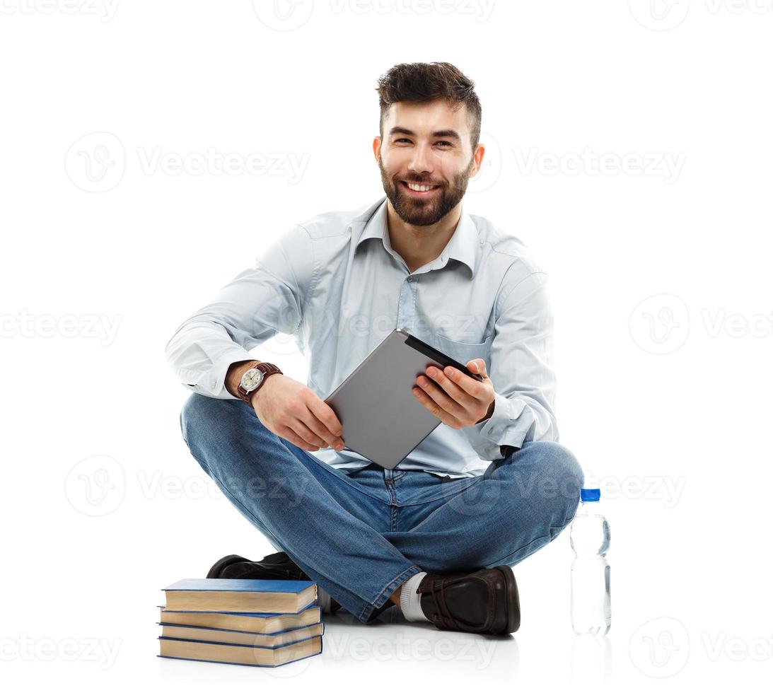 Young bearded smiling man holding a tablet with books and a bottle of water sitting on a white photo