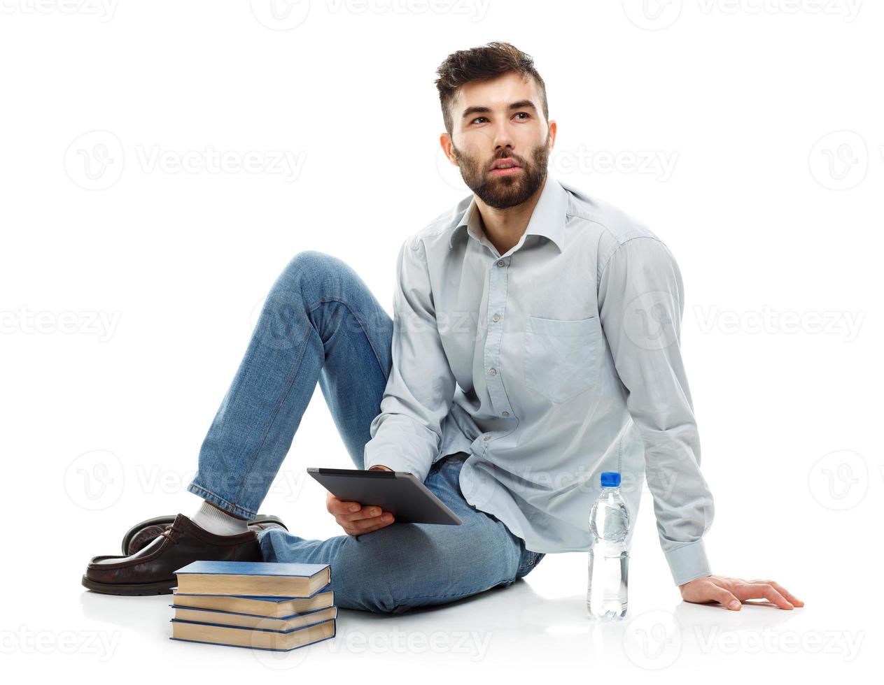 Young bearded man holding a tablet with books and a bottle of water sitting on a white photo