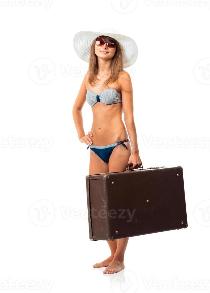 Full length portrait of a beautiful young woman posing in a bikini, hat and sunglasses with a suitcase in hand on white photo