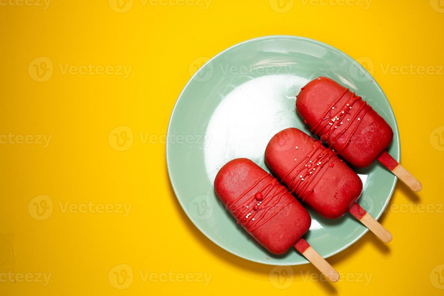 Cake in the form of ice cream on a stick among flowers, close-up. Cheesecake on a stick. Ice cream in glaze. Yellow background, top view photo