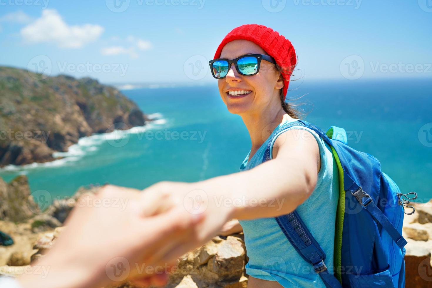 seguir yo - contento joven mujer en un rojo sombrero y con un mochila detrás su espalda tracción tipo mano. mano en mano caminando a el Oceano costa foto