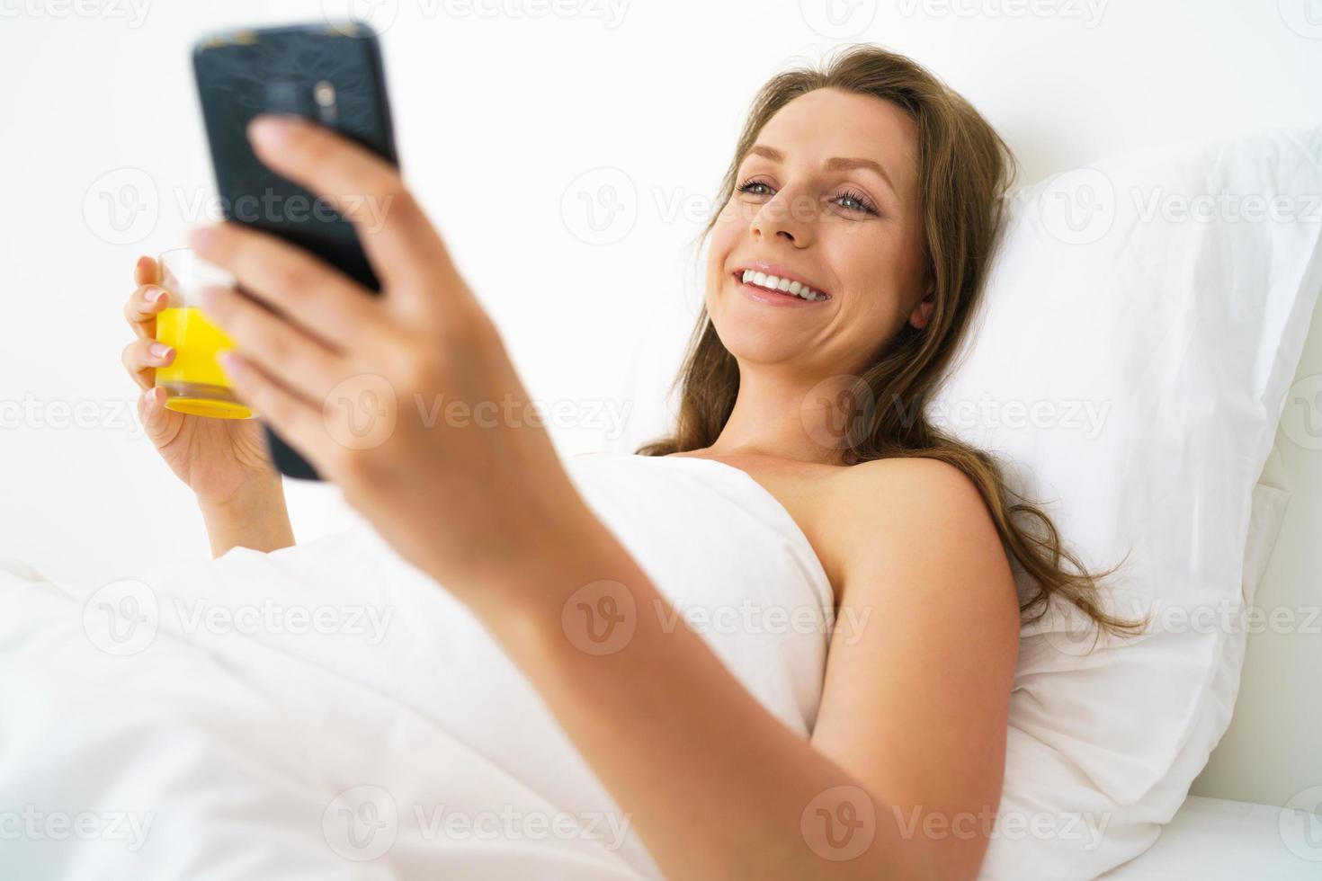 Cute woman checks the smartphone and drinks orange juice in bed in the morning photo