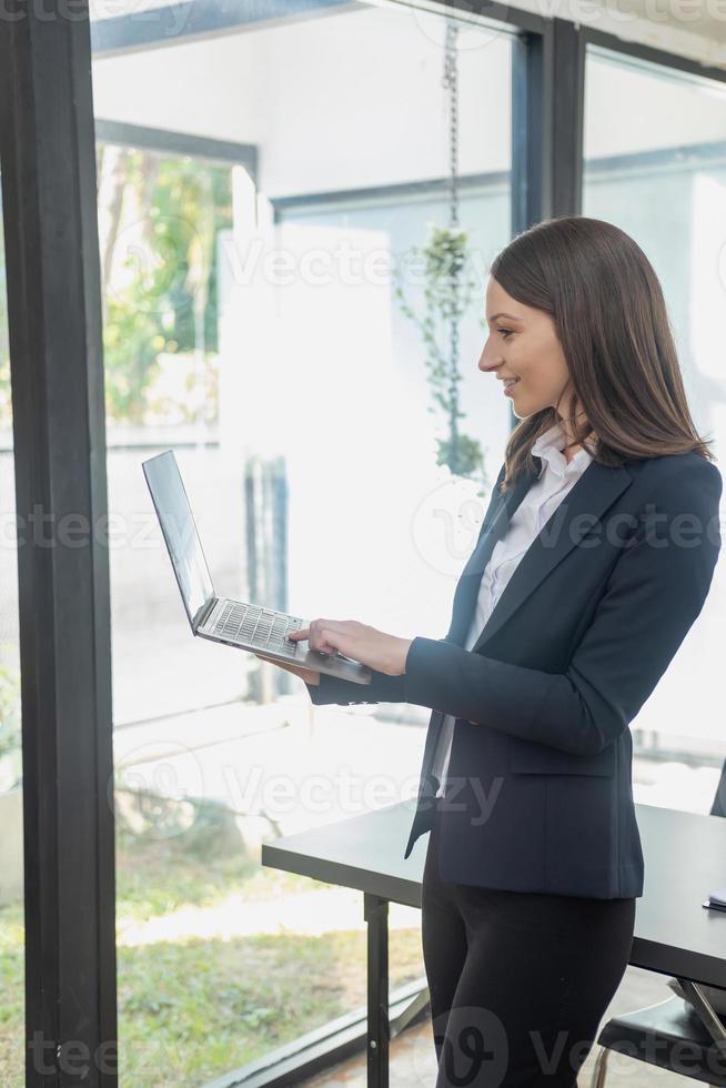 A stylish white businesswoman in a black suit standing inside the office in hands with a mobile phone, tablet, and laptop waiting for customers to contact, business people concept. photo