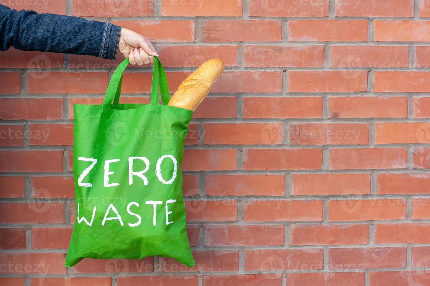 Eco bag with the inscription zero waste in hand on a background of a brick wall with copy space. French bread sticks out of the bag. photo
