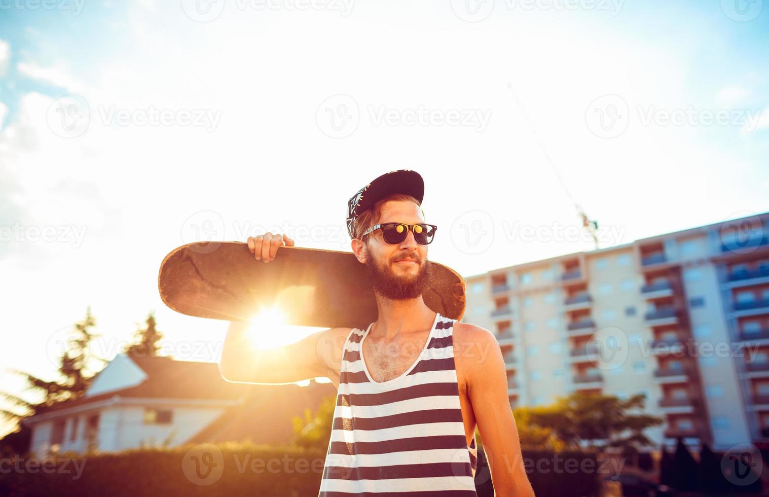 Man in sunglasses with a skateboard on a street in the city at sunset light photo