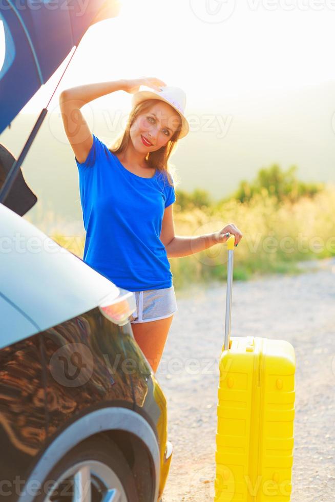 Young woman with a yellow suitcase standing near the trunk of a car photo