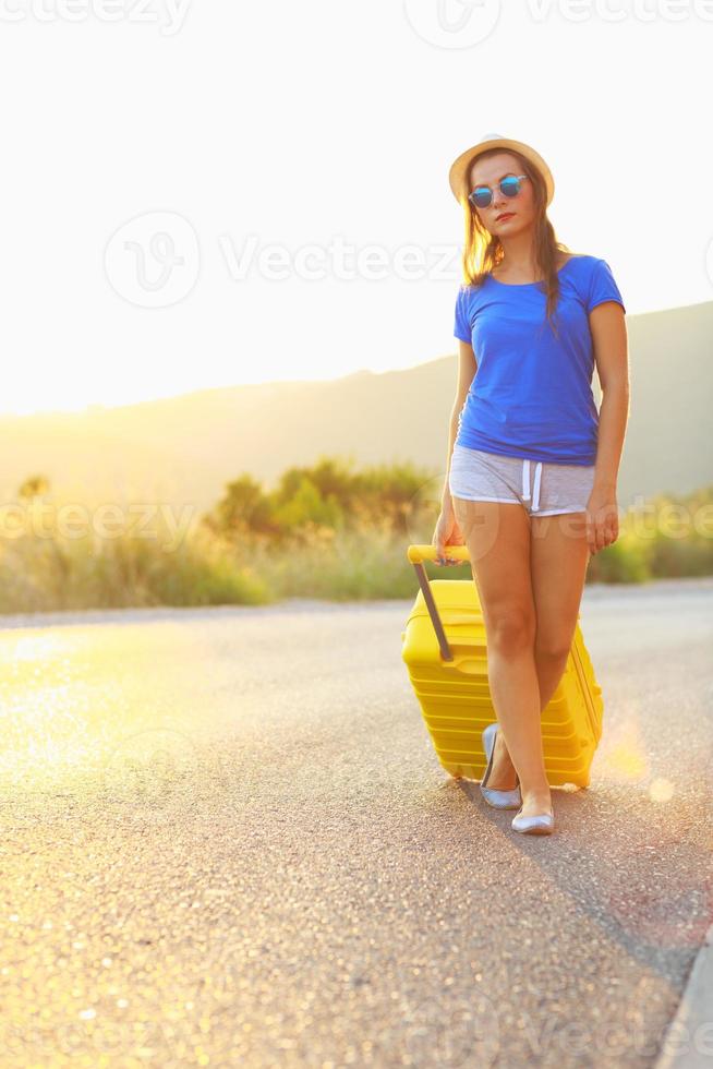 Attractive young woman with a yellow suitcase is traveling on the road hitchhiking photo