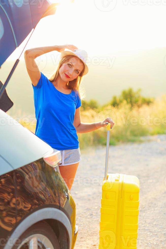 Woman with a yellow suitcase standing near the trunk of a car parked on the roadside photo