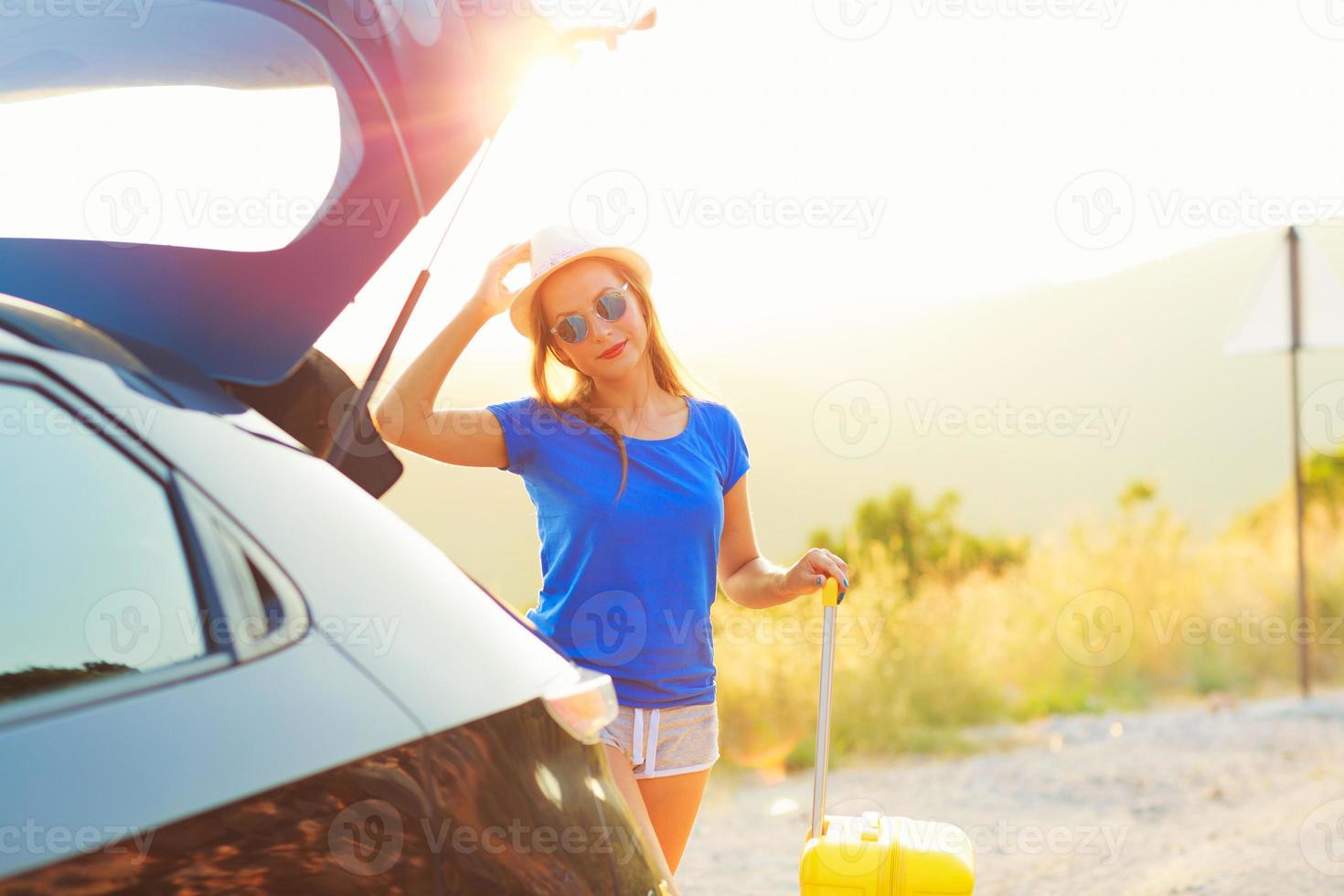 Woman with a yellow suitcase standing near the trunk of a car parked on the roadside photo