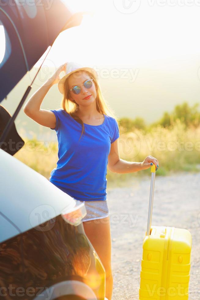 Woman with a yellow suitcase standing near the trunk of a car parked on the roadside photo