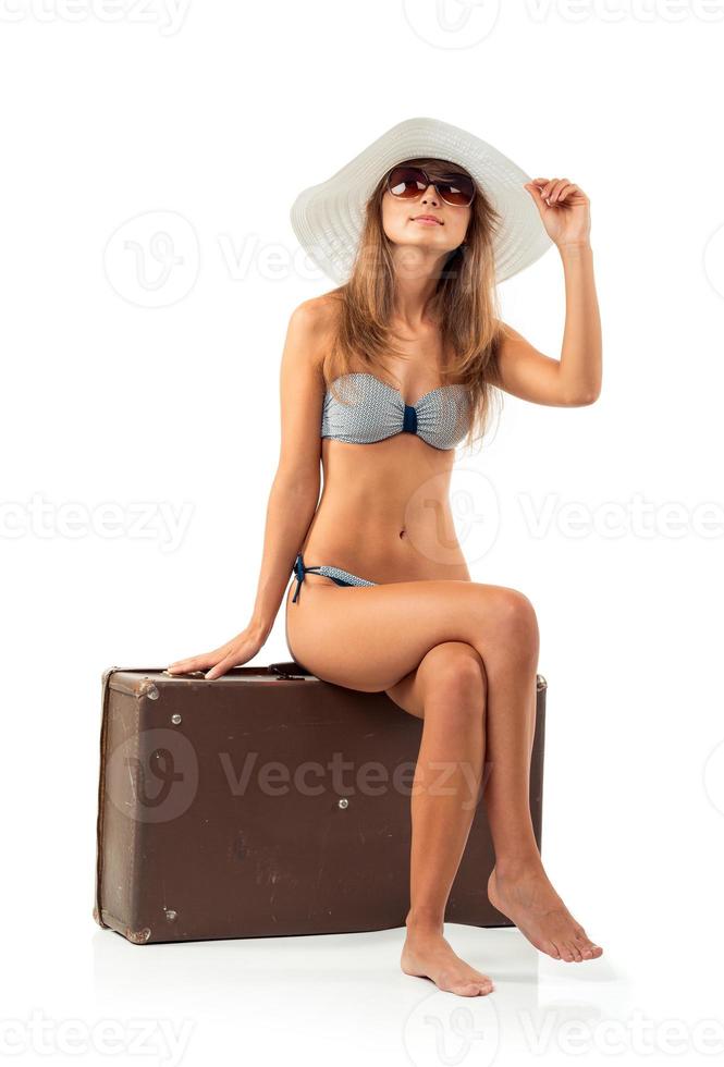 Full length portrait of a beautiful young woman posing in a bikini sitting on a suitcase on a white photo