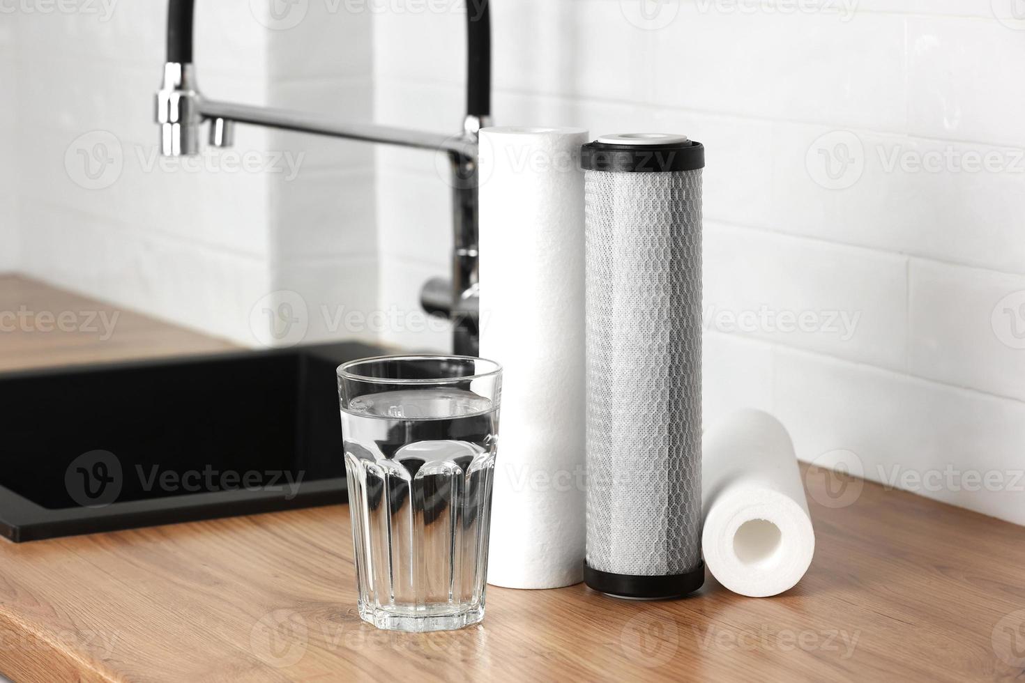 A glass of clean fresh water and set of filter cartridges on wooden table in a kitchen interior. Installation of reverse osmosis water purification system. Concept Household filtration system. photo
