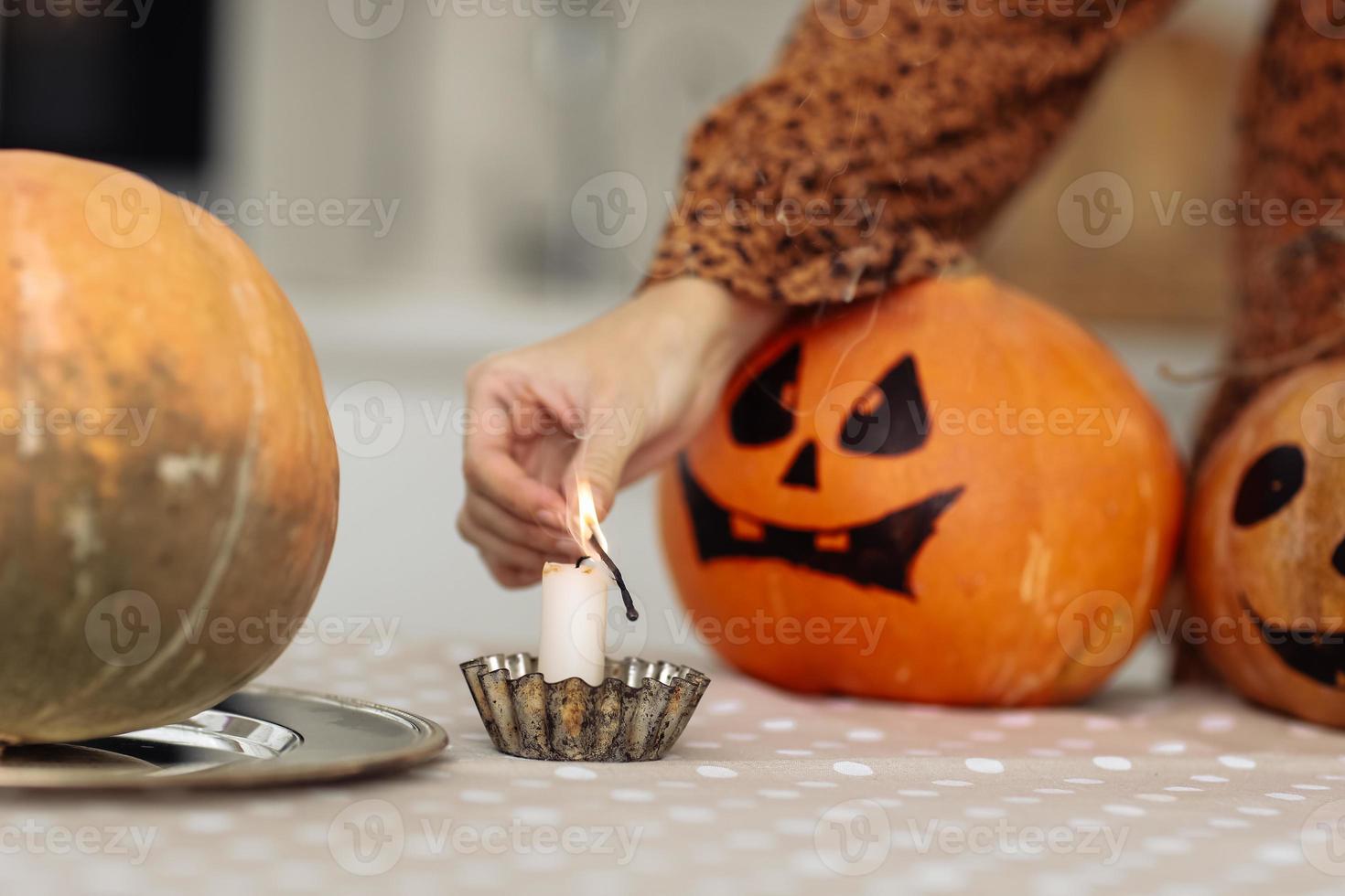 holidays and leisure concept - woman's hand with matches lighting candles at home on halloween. painted pumpkins on a background. horror theme and Hallowe'en. selective focus. photo