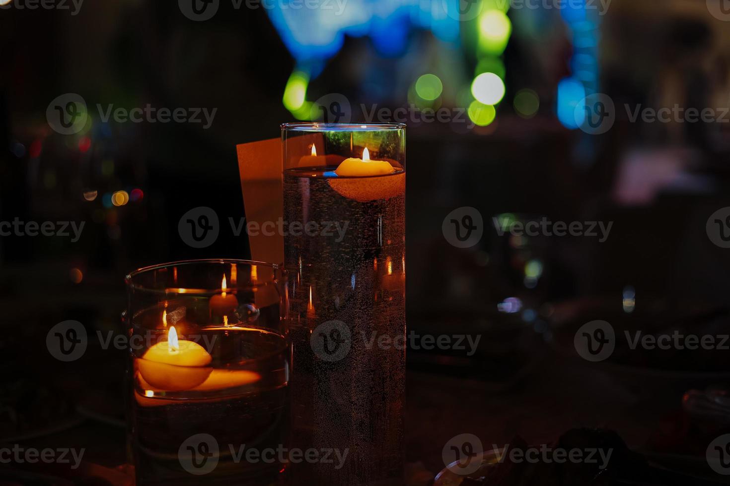 Banquet table decorated with burning candles in glass vases in restaurant hall. In the background party with silhouettes of people dancing on the dance floor with disco lights glowing searchlight photo