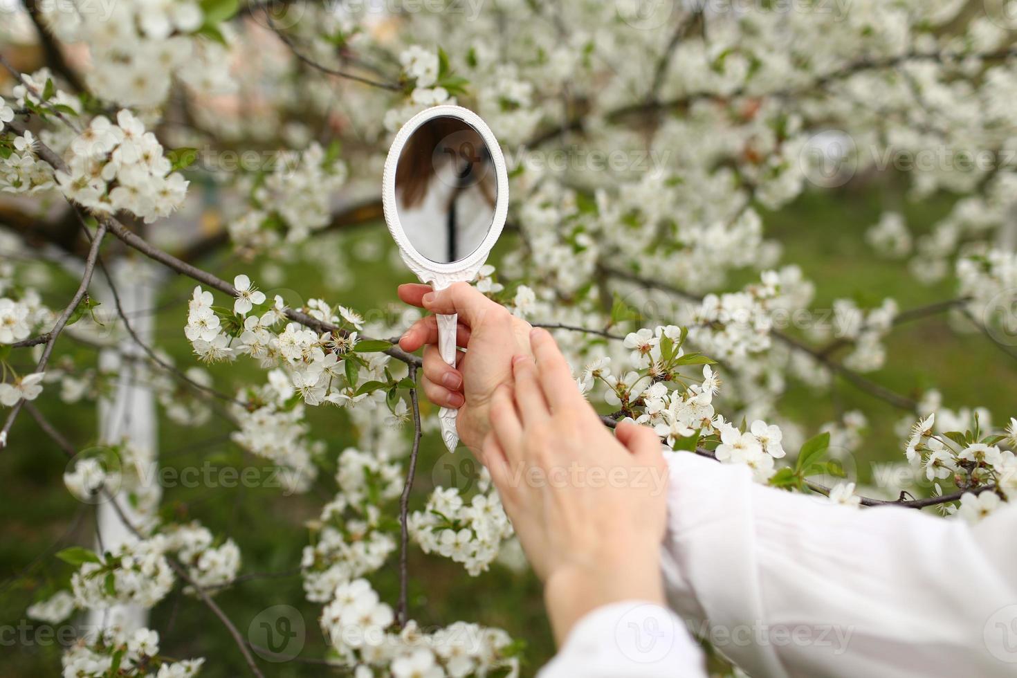 Outdoor close up of young female hand holding small retro mirror in blooming garden on spring day. Model looking in little mirror, posing in street, near flowering trees. Female fashion concept photo