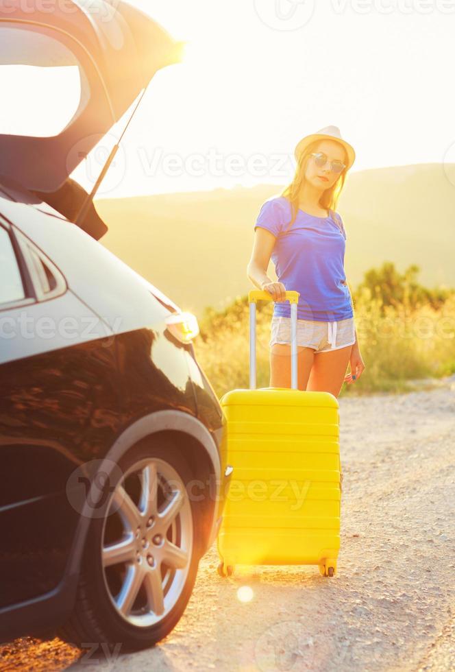 Woman with a yellow suitcase standing near the trunk of a car parked on the roadside photo
