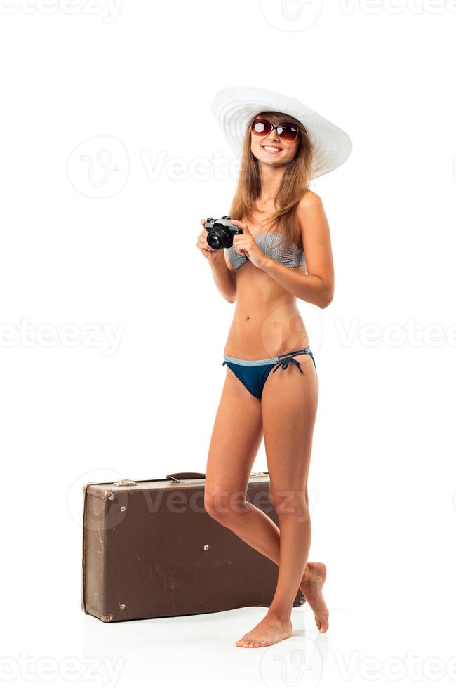lleno longitud retrato de un hermosa joven mujer posando en un bikini con un cámara en manos y maleta en blanco foto