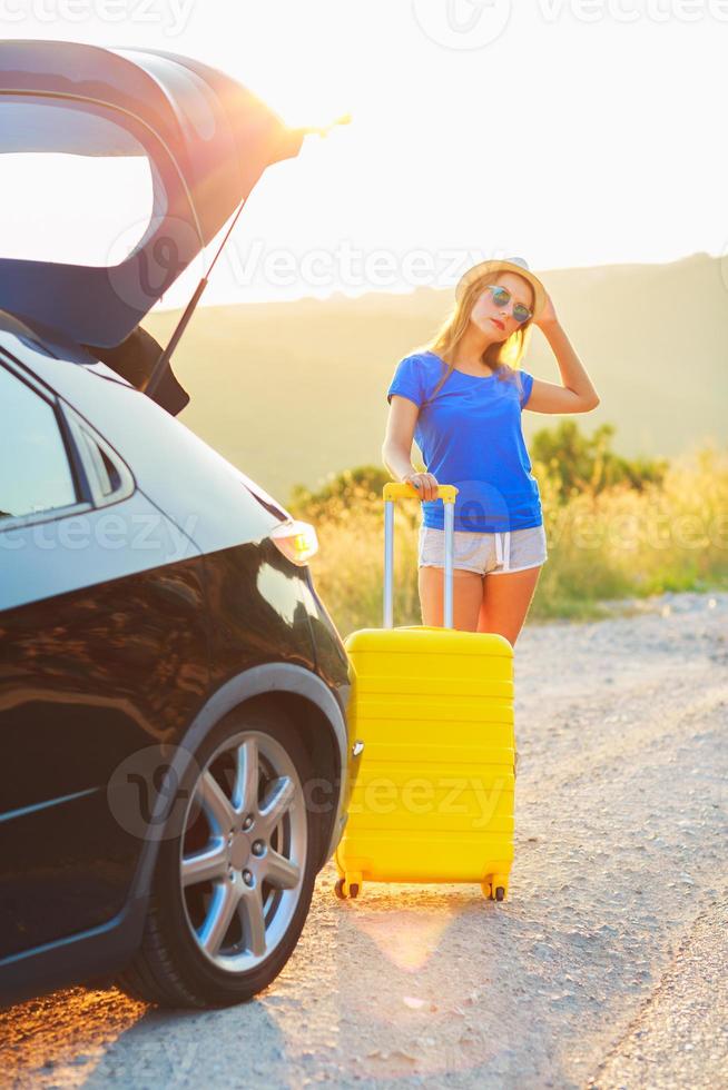 Young woman with a yellow suitcase standing near the trunk of a car photo