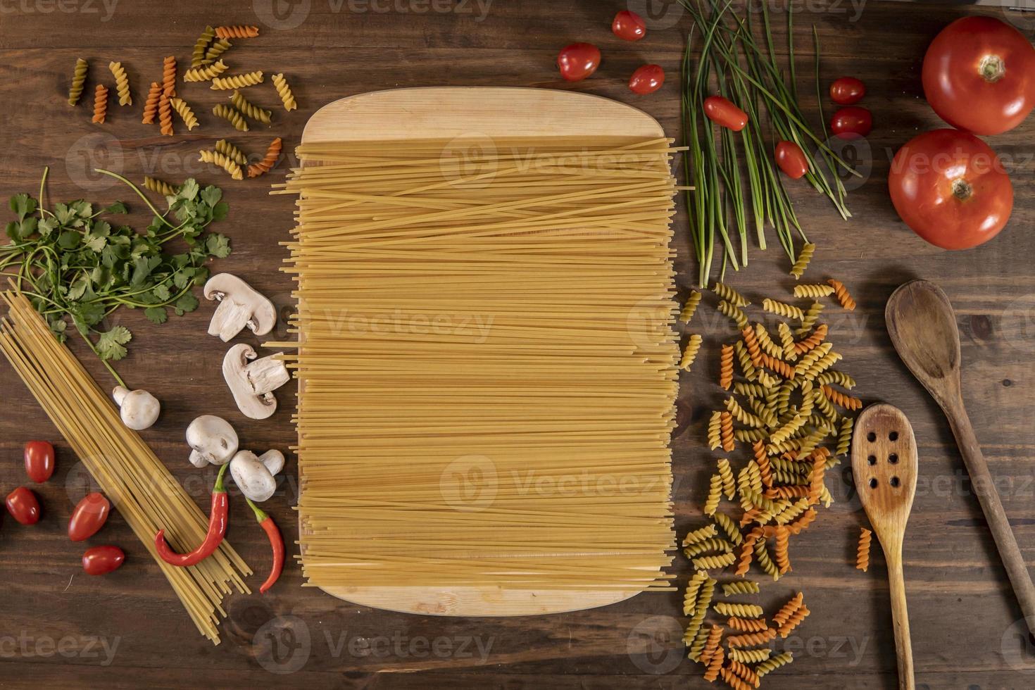 Flat lay of vegetables and pasta and a chopping table over a wooden table photo