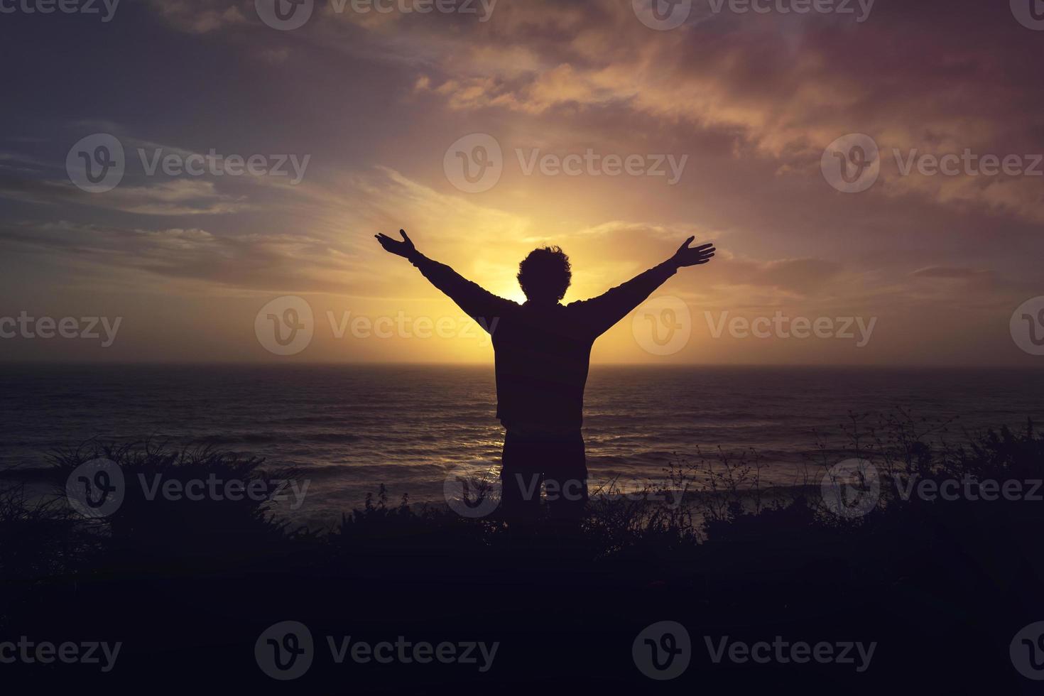 Silhouette Of A Man Watching orange Sunset on cliff over the sea with open arms photo