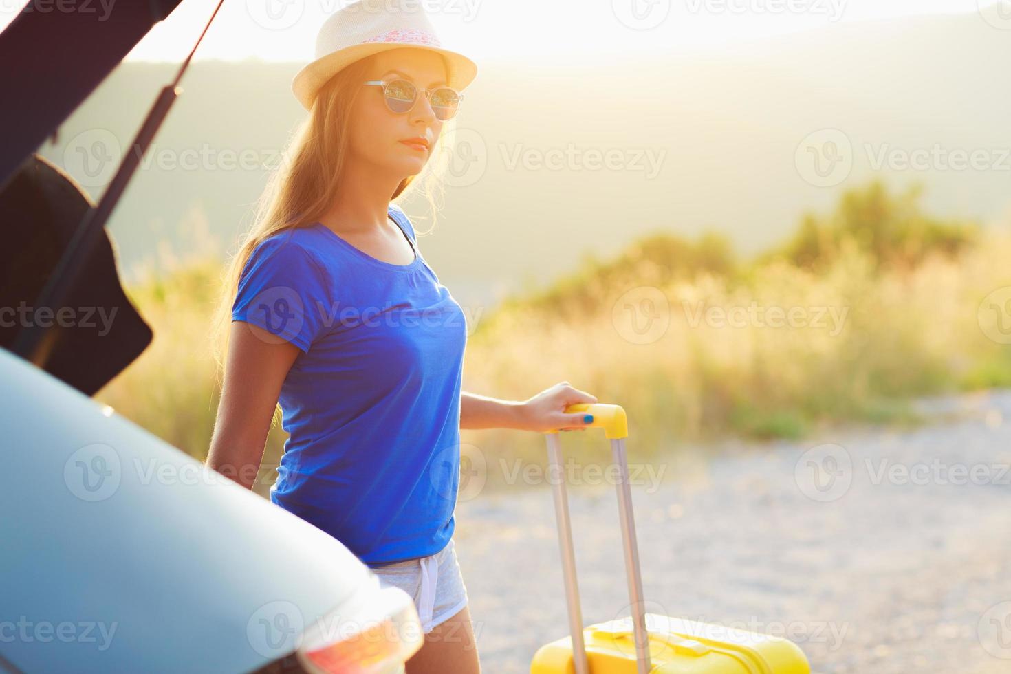Woman with a yellow suitcase standing near the trunk of a car parked on the roadside photo
