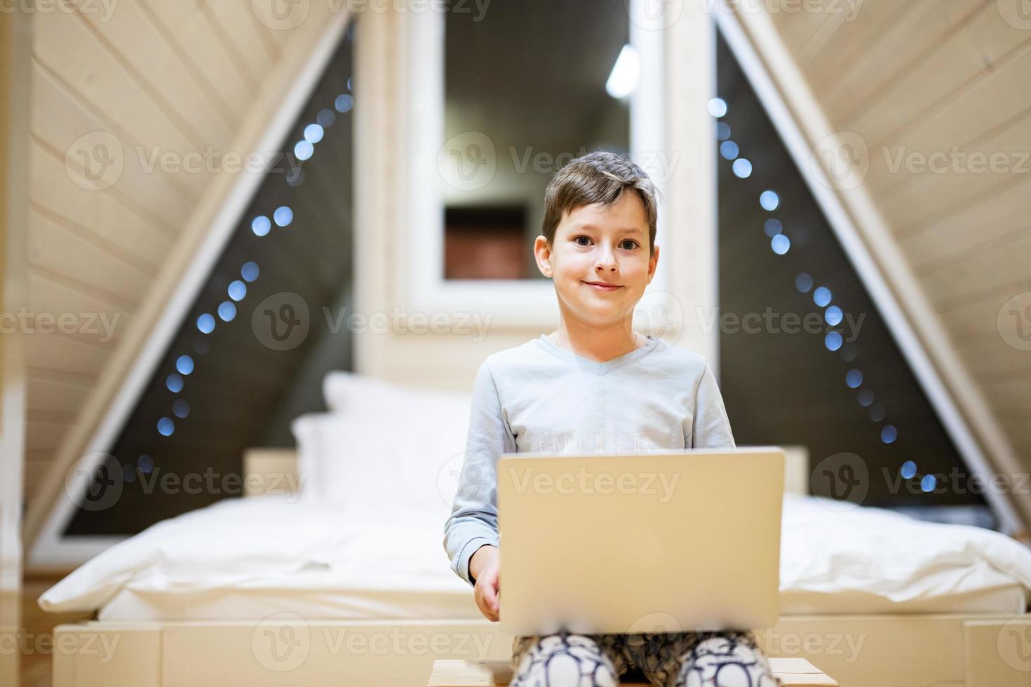 Boy in pajama sit with laptop at wooden cabin home. Concept of childhood, leisure activity, happiness. photo