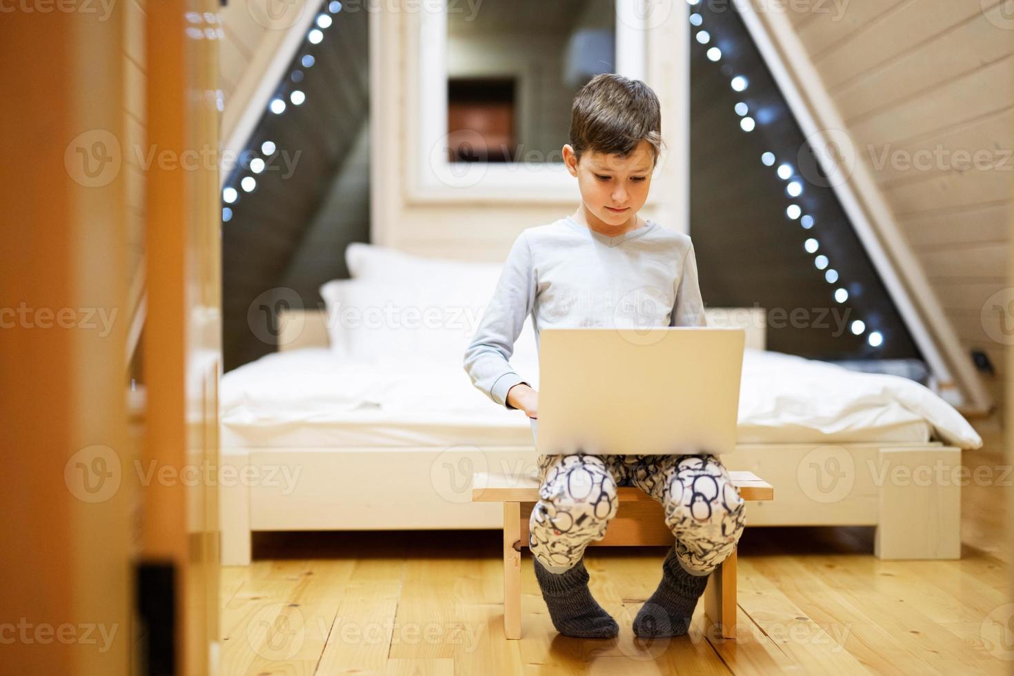 Boy in pajama sit with laptop at wooden cabin home. Concept of childhood, leisure activity, happiness. photo
