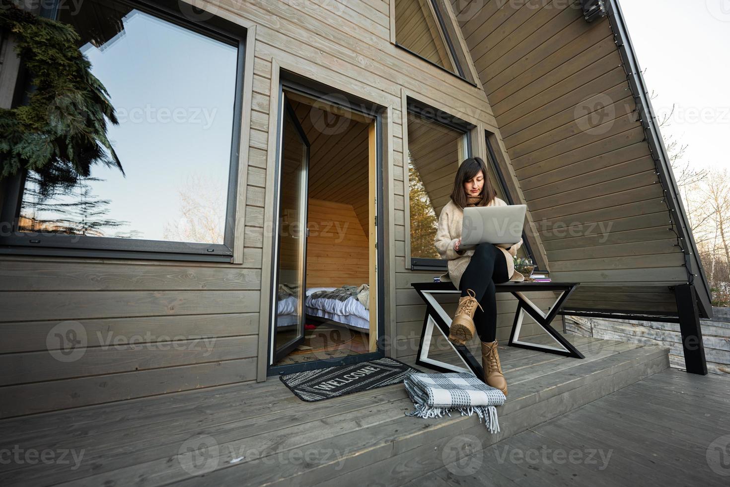 Remote work and escaping to nature concept. Woman works on laptop against tiny cabin house. photo