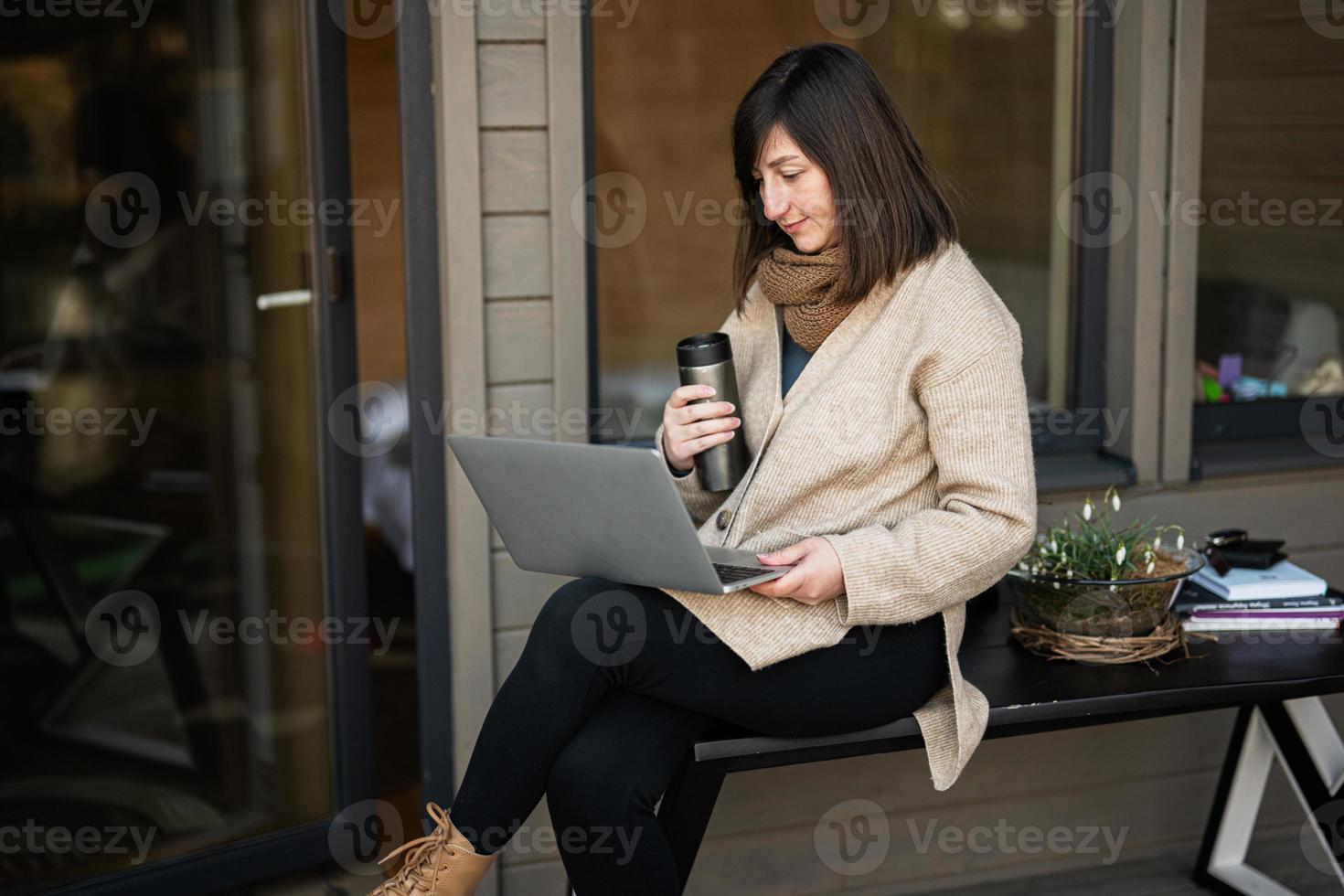 Remote work and escaping to nature concept. Woman works on laptop and drink tea from thermos against tiny cabin house. photo