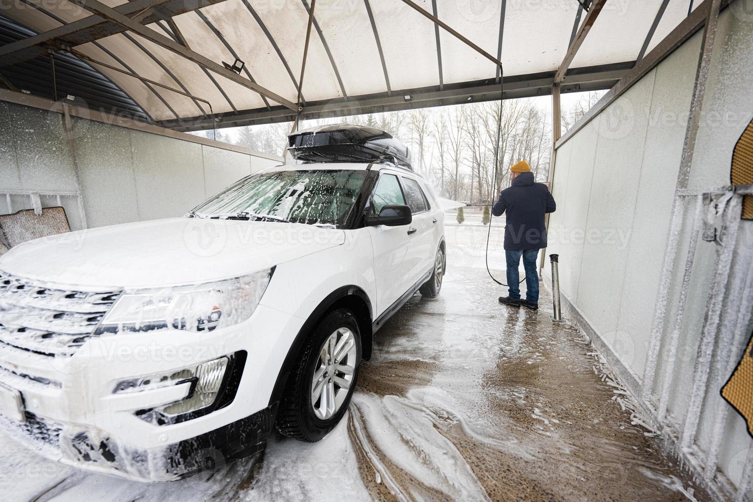 hombre lavando agua a alta presión coche suv americano con portaequipajes en autoservicio de lavado en clima frío. foto