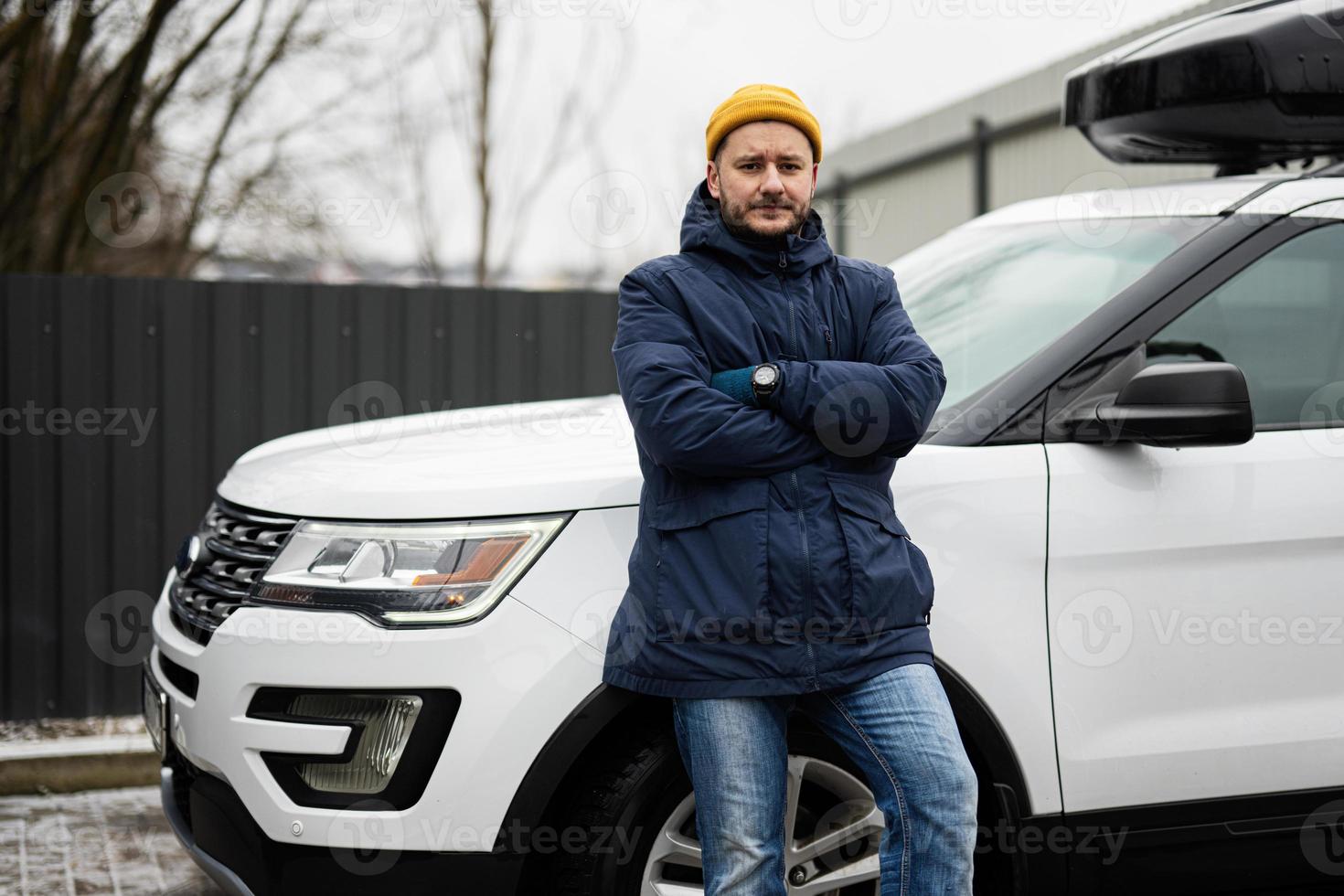 Man driver wear jacket and yellow hat against his american SUV car with roof rack in cold weather. photo