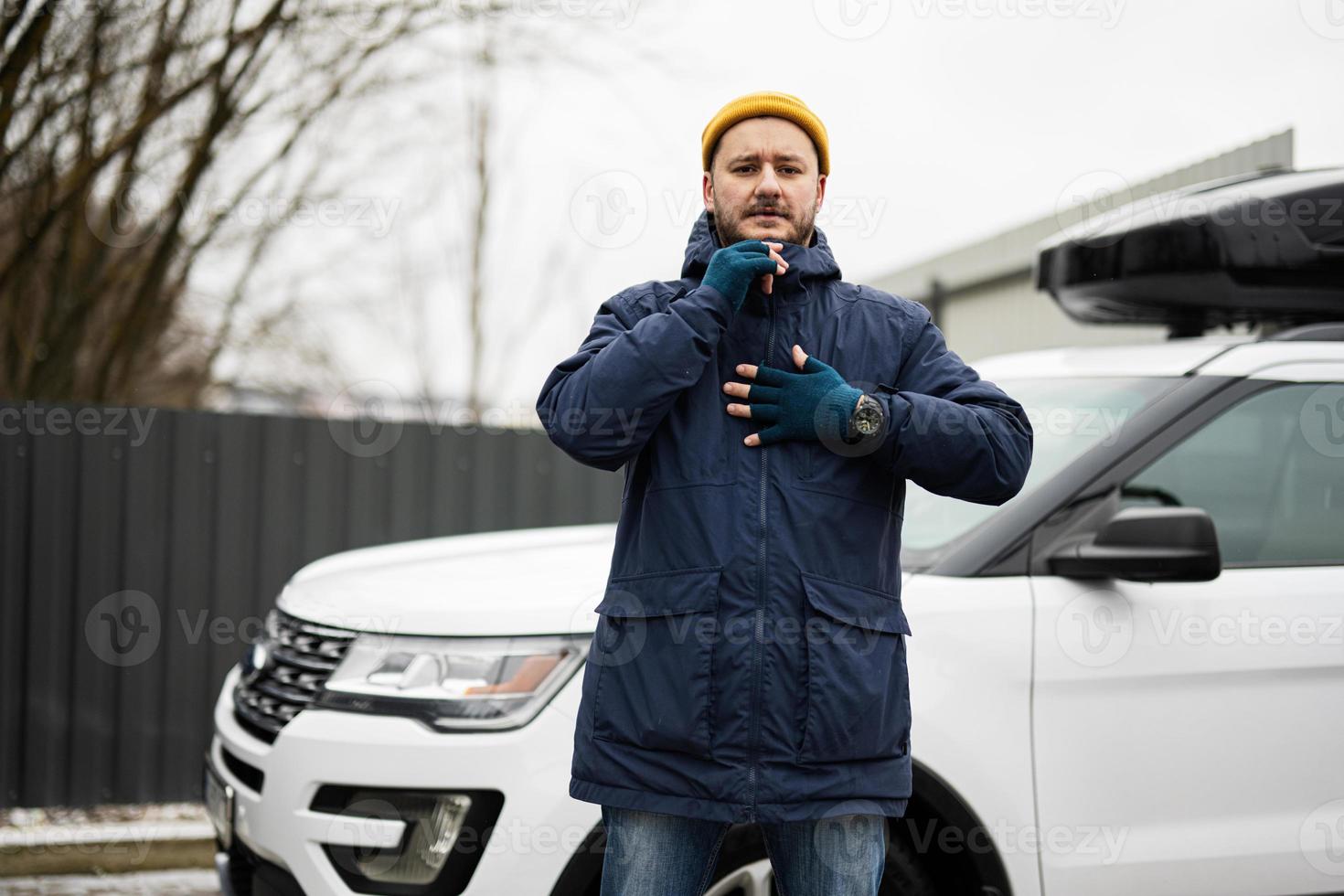 Man driver wear jacket and yellow hat against his american SUV car with roof rack in cold weather. photo