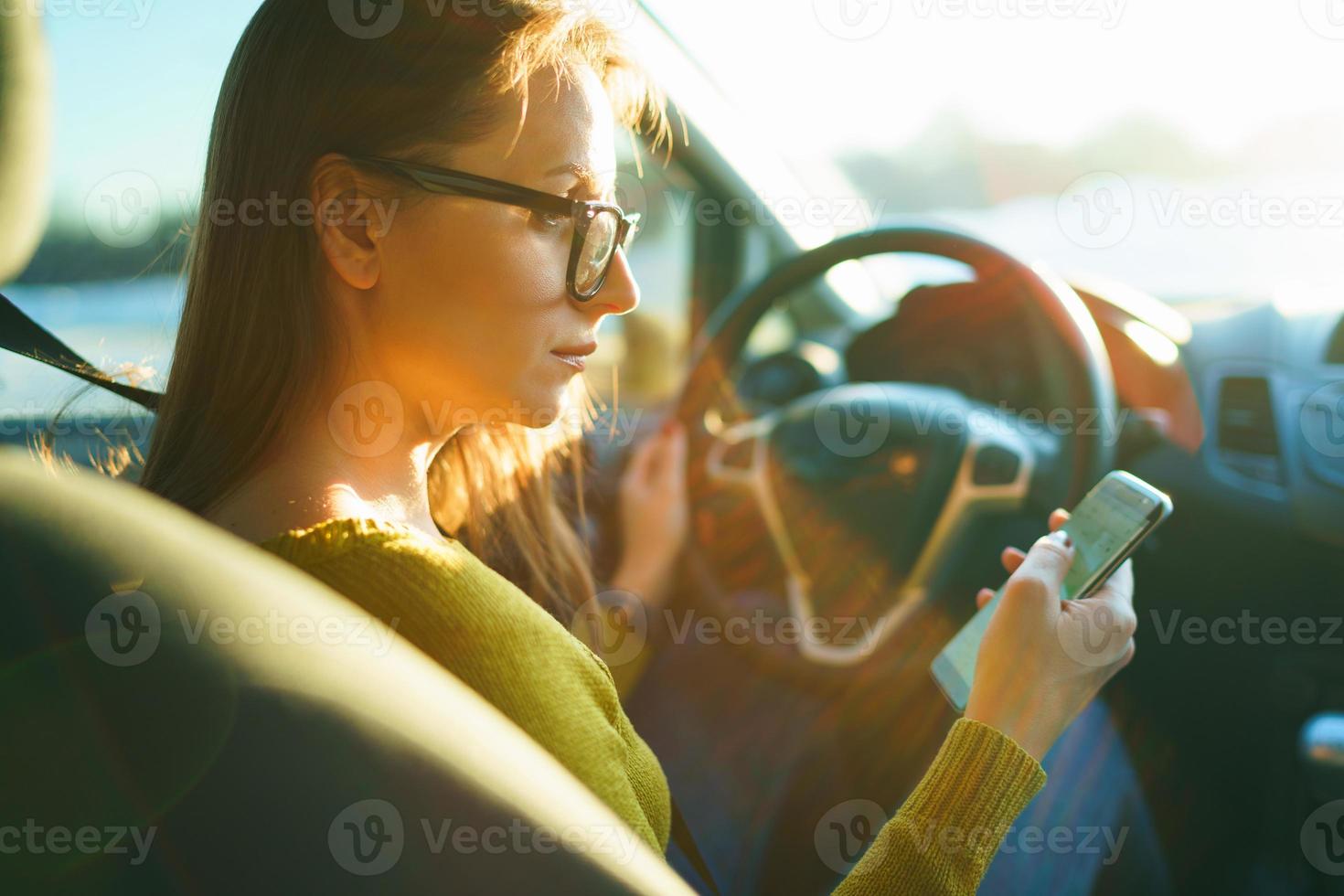 Woman in glasses uses a smartphone while driving a car at sunset photo