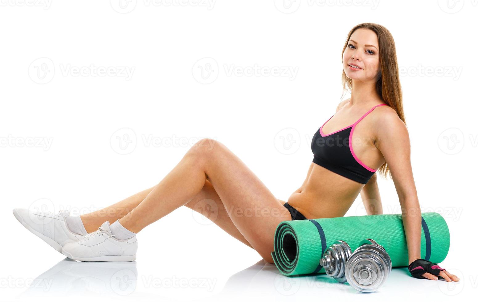 Beautiful athletic woman with dumbbells, mat for fitness, a bottle of water sitting on white photo