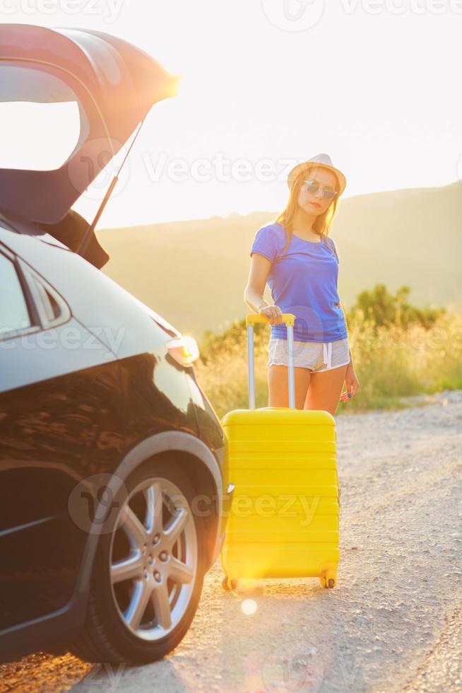 Woman with a yellow suitcase standing near the trunk of a car parked on the roadside photo