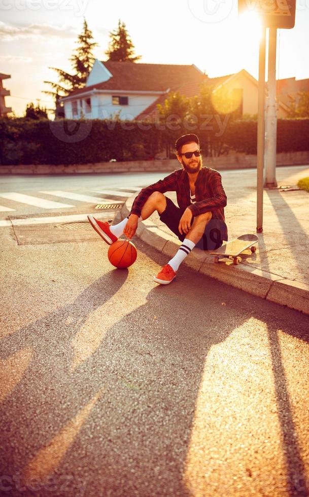 Man in sunglasses with a basketball and skateboard sitting on a city street at sunset light photo