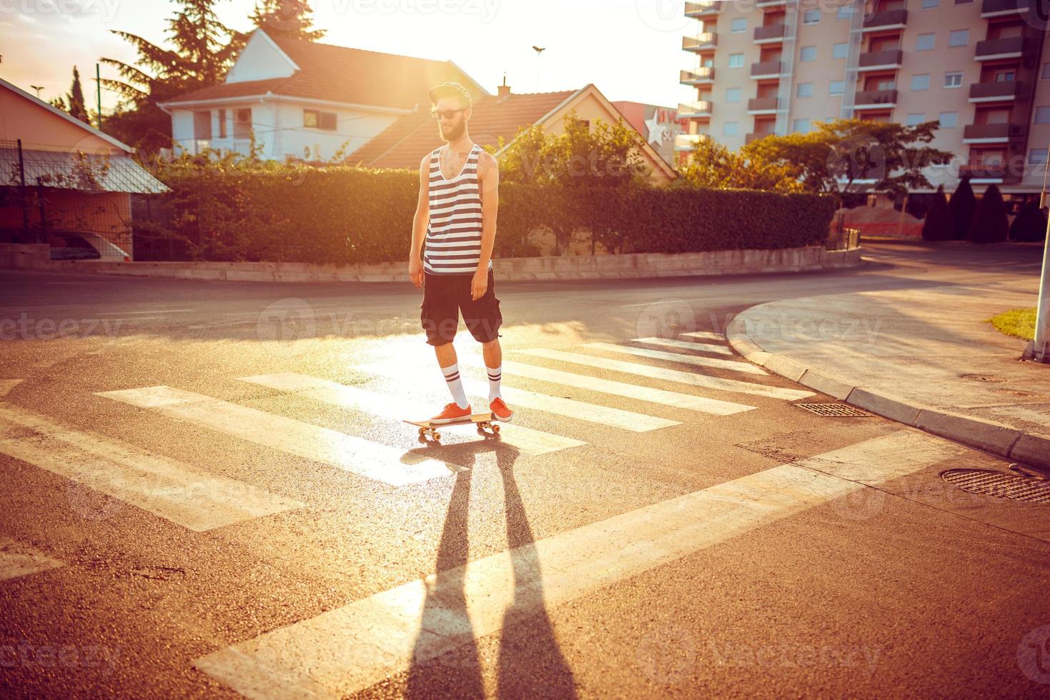 Young man in sunglasses with a skateboard on a street in the city at sunset light photo