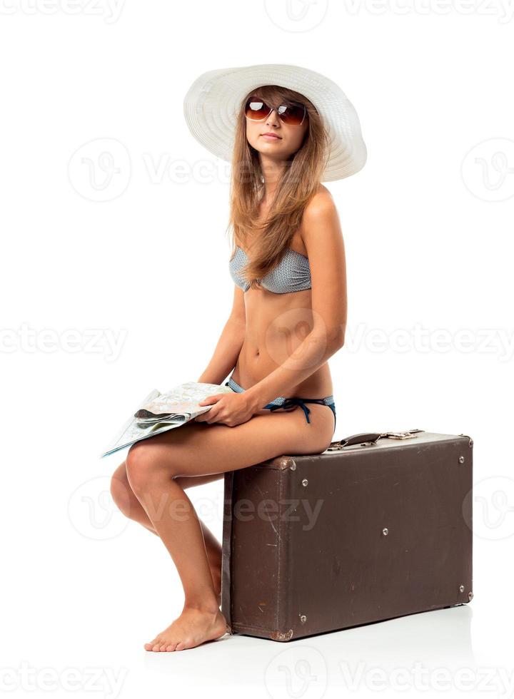 Full length portrait of a beautiful young woman posing in a bikini sitting on a suitcase with map on a white photo