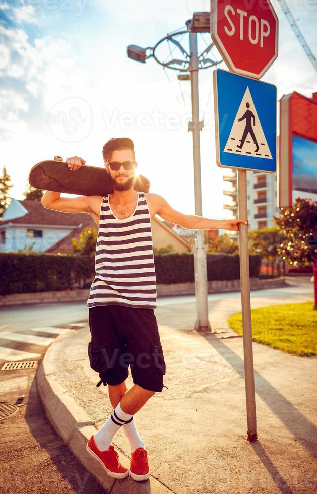 Young man in sunglasses with a skateboard on a street in the city at sunset light photo